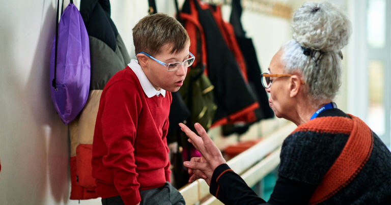A male-presenting student wearing glasses and a red sweater sits on a bench in front of a wall where backpacks and coats are hanging. In front of him sits his teacher, a female-presenting person with glasses, grey hair, and a black and orange sweatshirt. They are talking to the student.